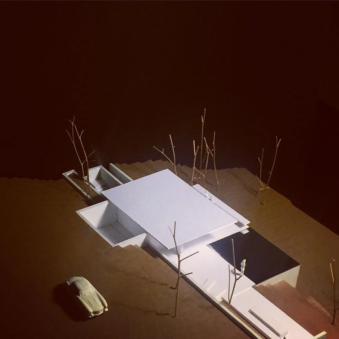 White box model with a dark roof lit dramatically over a sandy ground with twig trees